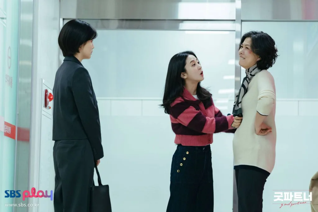 A woman in dark suit is watching another woman dressed in a white blouse while she is adressign her in anger tones. Another woman in pink and black blouse is trying to calm the woman in the white blouse.  A still from the Good Partner drama.