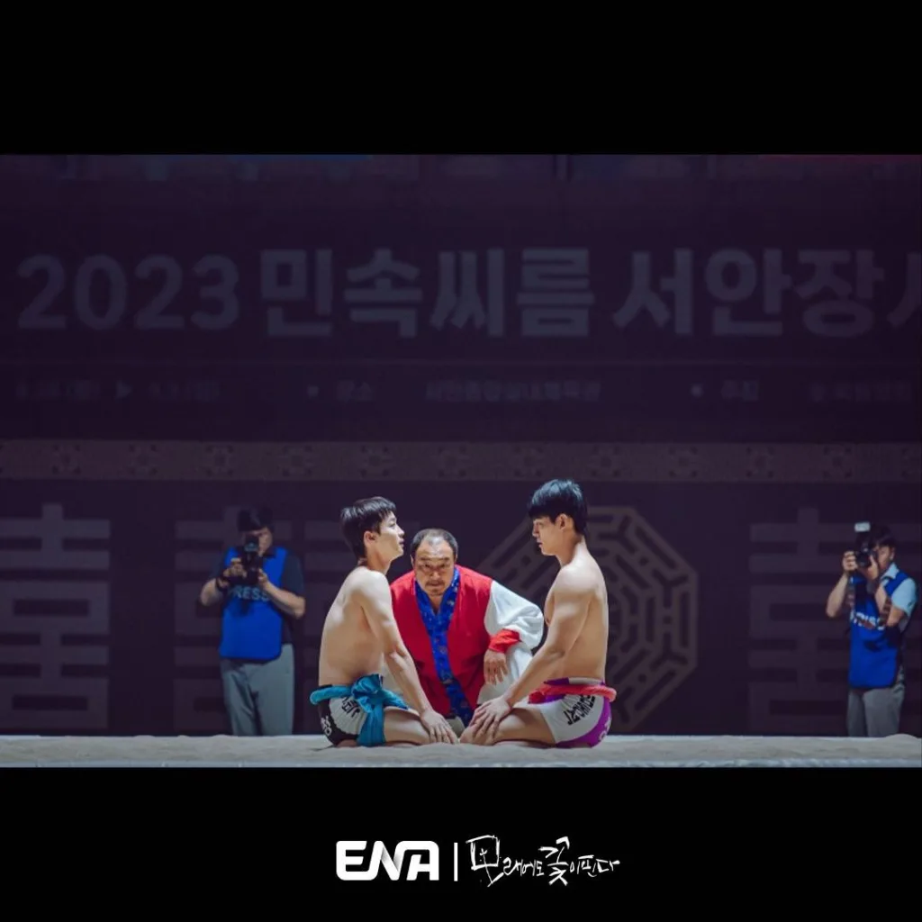 Two athletes of  ssireum, a traditional Korean wrestling sport are sitting on the wresting pit facing each other. A still image from a sports Korean drama