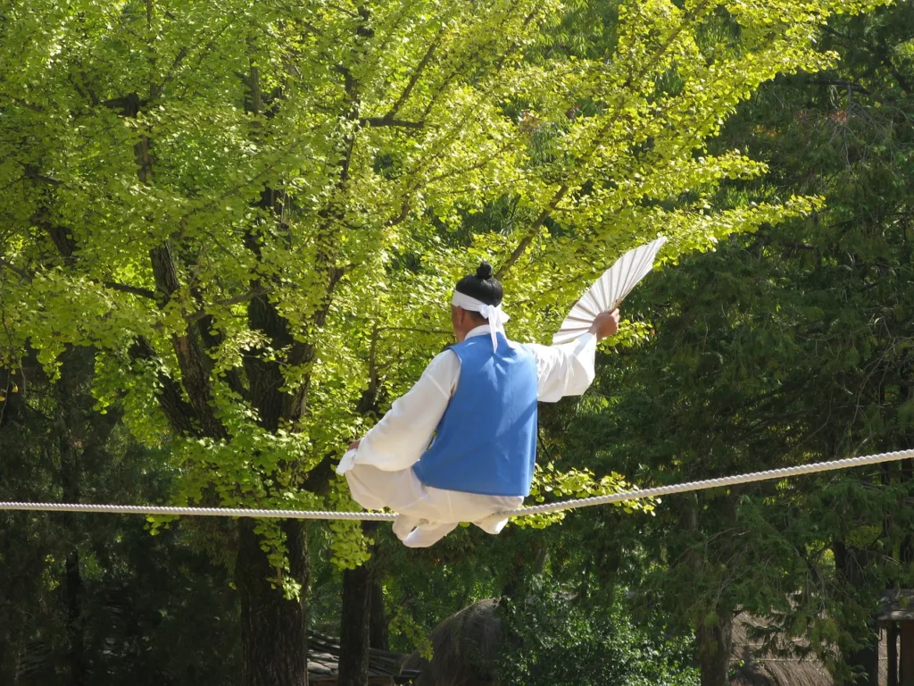 a man wearing a traditional Korean male  dress is balancing on a rope holding a fan during filming of Korean Dramas