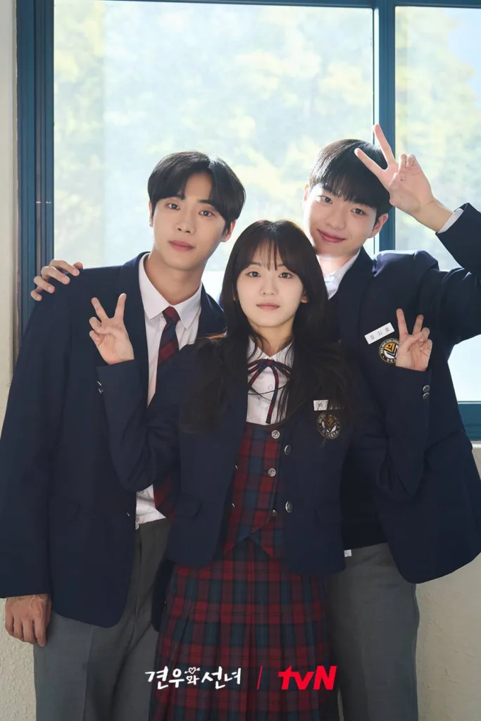 Promotional photo for the Korean drama "Gyeonwoo and Seon-nyeo" (tvN). Three young actors in school uniforms pose together indoors near a large window with green trees visible outside. A young woman stands in front, wearing a navy blazer over a red-and-black plaid dress with a bow tie; she looks directly at the camera with a soft smile. Behind her, two young men flank her — one on the left makes a peace sign with his right hand, while the other on the right smiles broadly, making peace signs with both hands. All three wear matching navy blazers with school badges and name tags. The atmosphere is cheerful and youthful.