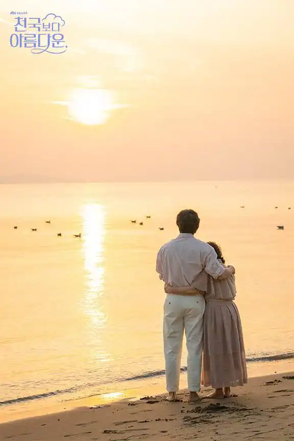 . A man and woman stand barefoot on a sandy beach at sunset, seen from behind as they gaze out over the calm ocean. The man, dressed in a light pink button-down shirt and white pants, has his arm wrapped gently around the woman, who wears a flowing mauve dress. Their heads are close together, sharing a moment of quiet connection as the golden sun dips toward the horizon, casting shimmering reflections across the water. Small birds float peacefully on the surface nearby. In the top left corner, the title “천국보다 아름다운” is written in elegant blue script with a cloud motif. The mood is tender, nostalgic, and deeply romantic — suggesting a love story that feels transcendent, healing, and worth cherishing. This is a promotional poster from the Korean dramas Heavenly ever after. One of the best drama series for self-care.
