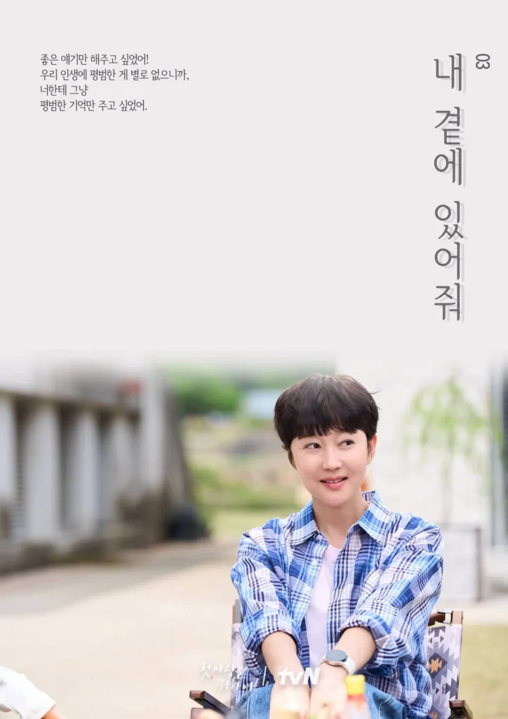 A woman with short dark hair and a soft, contented smile sits outdoors in a director’s chair, wearing a blue-and-white plaid shirt over a white tee. She looks off to the side with a gentle, reflective expression, as if lost in a fond memory or quiet hope. The background is softly blurred — showing a rustic outdoor path, greenery, and hints of a building — evoking calm, everyday beauty. Korean text on the left reads: “좋은 얘기만 해주고 싶었어! 우리 인생에 평범한 게 별로 없으니까, 너한테 그냥 평범한 기억만 주고 싶었어.” (“I only wanted to tell you good stories! Since ordinary things are rare in our lives, I just wanted to give you simple, ordinary memories.”). On the right, vertical text says: “내 곁에 있어줘” (“Stay by my side”). The show’s title and “tvN” logo appear subtly at the bottom. The mood is tender, nostalgic, and deeply human — suggesting a story about healing, connection, and finding peace in small moments. A poster from the Korean drama Love, Take Two.