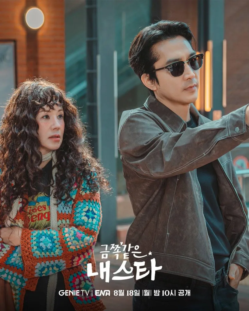 A woman with voluminous curly hair and freckles stands on the left, arms crossed, wearing a colorful crocheted cardigan over a graphic tee — her expression is slightly wary or surprised as she glances toward the man beside her. On the right, a man in sleek sunglasses and a dark leather jacket points confidently ahead, one hand in his pocket, exuding cool charisma. They stand side by side against a modern urban backdrop with warm brick walls and glowing vertical lights — suggesting a stylish city setting. The title “금쪽같은 내 스타” is displayed in bold white lettering at the bottom, with “GENIE TV | ENA 8월 18일 [월] 밤 10시 공개” beneath it. The mood is playful, stylish, and full of chemistry — hinting at a rom-com where opposites clash, then charm each other into love.Promotional poster for the Genie TV Original drama “금쪽같은 내 스타” (Geumjjokgat-eun Nae Seuta /My Troublesome Star.