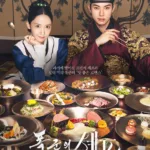 A young woman in a traditional blue-and-white hanbok with an elegant updo sits beside a man in a luxurious maroon royal robe adorned with golden dragon embroidery and a black gat (traditional hat). Both gaze confidently at the viewer — she rests her chin on her hand with a knowing smile, while he holds chopsticks mid-air, as if about to taste one of the many dishes before them. Spread across the table is an extravagant feast: modern gourmet plates featuring seared steak, colorful vegetable arrangements, delicate pastries, and artistic plating — all served on gleaming silverware against a backdrop of ornate Korean folding screens with grapevine patterns. Korean text reads: “과거에 떨어진 프렌치 셰프와 절대 미각 폭군의 ‘맛 좋은 로맨스’” (“A French chef who fell into the past and an absolute taste tyrant — a delicious romance”). The title “폭군의 쉐프” is displayed in bold white brushstroke lettering at the bottom, with “tvN 토일 드라마” and “TVING 8월 23일 공개” beneath it. The mood is lavish, romantic, and mouthwateringly stylish — promising a historical fantasy where culinary passion meets royal power. Promotional poster for the tvN Saturday-Saturday drama “폭군의 쉐프” (Pokgun-ui Syeopeu / The Tyrant’s Chef), premiering August 23 on TVING. Part of the best food-themed k-dramas.