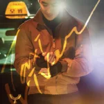 The official poster for K-drama Taxi Driver 3, a top action series in the november K-drama lineup, showing a man in a bomber jacket smiling softly under city lights with his taxi behind him.