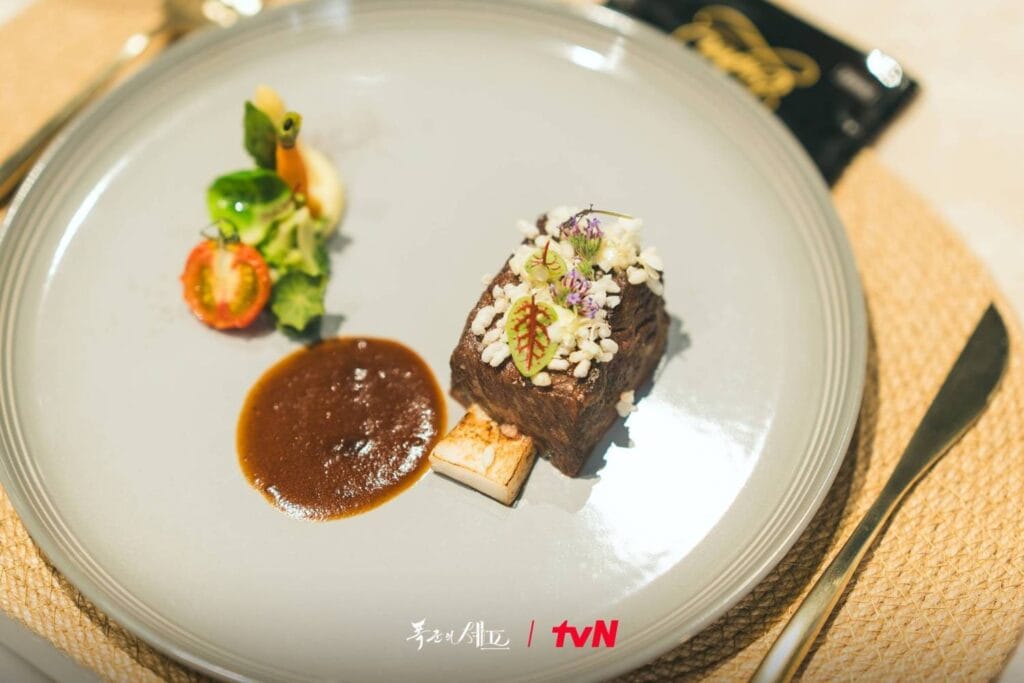 Gourmet dish from Bon Appétit, Your Majesty— seared beef with edible flowers, sauce, and fresh veggies, plated with fine-dining precision.