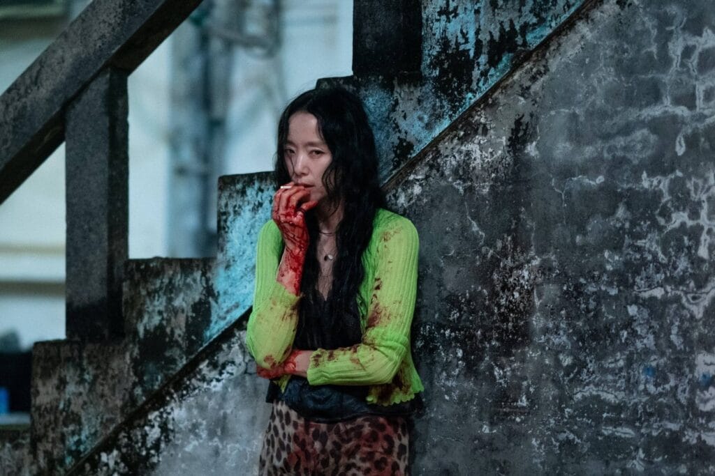 A blood-stained woman in a green cardigan leans against a decaying stairwell, her face empty with shock or grief.  A still from The Price of Confession.