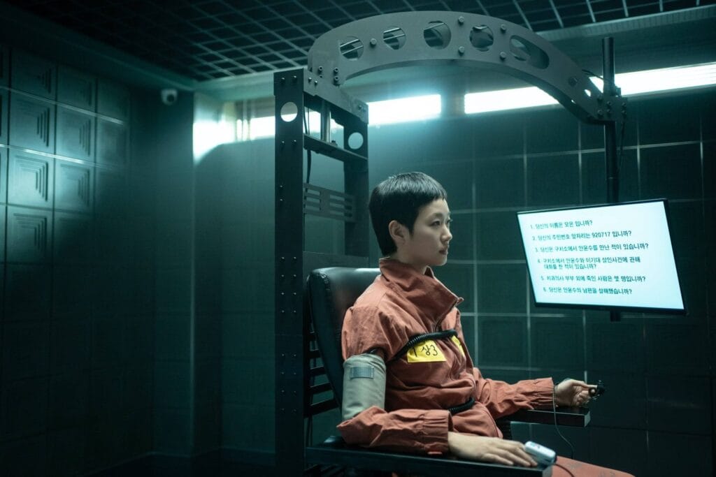 A woman in an orange prison jumpsuit sits strapped in a chair during a high-tech interrogation, answering questions on a glowing screen.  A still from The Price of Confession.