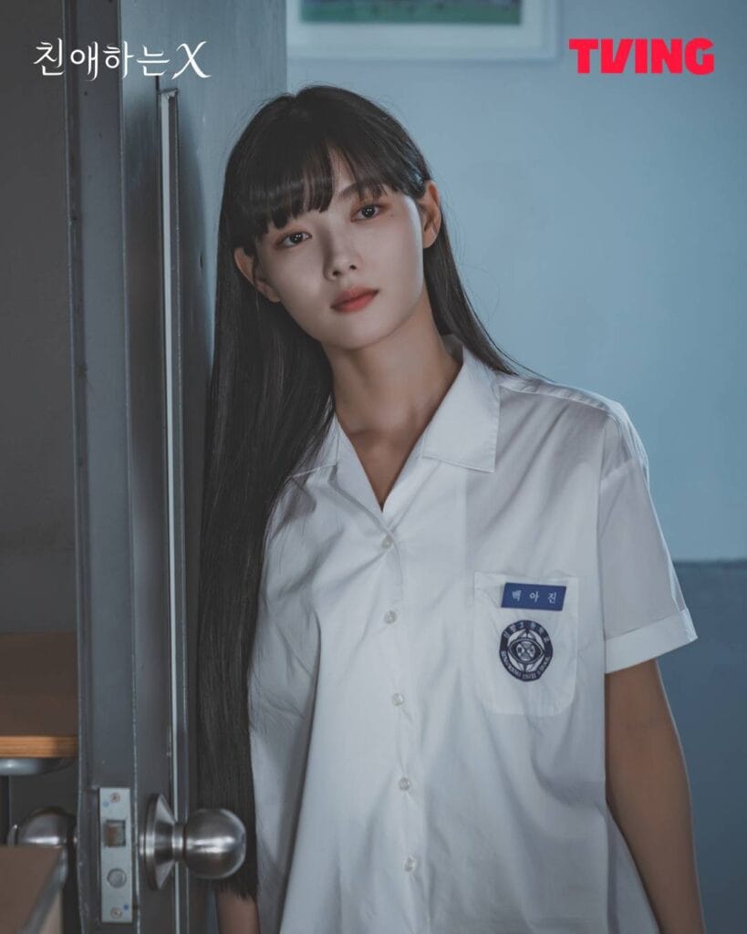 A young woman with long dark hair and bangs stands in a school hallway, leaning slightly against a metal doorframe. She wears a crisp white short-sleeved school uniform shirt with a name tag reading “백아진” (Baek Ah-jin) and a circular school emblem on the chest pocket.