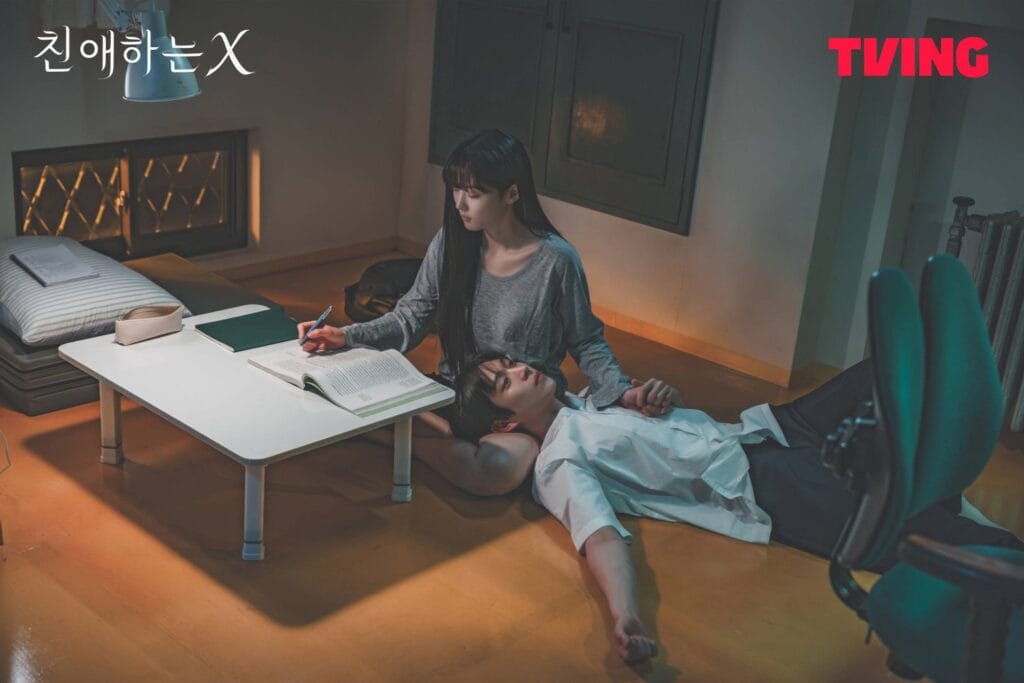A young woman with long dark hair sits on the floor beside a small white desk, gently holding the hand of a young man lying down with his head resting in her lap. A still  image from Dear X Korean Drama.