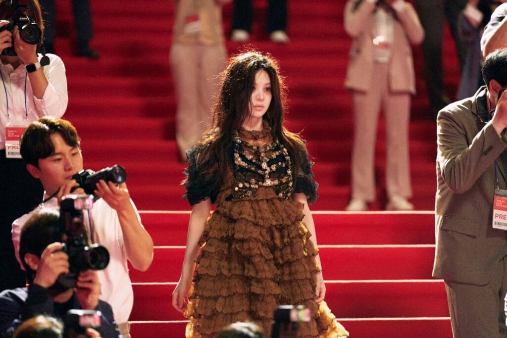 Go Youn-Jung with long, tousled dark hair walks down a grand red-carpeted staircase at the Tokyo Film Festival, her expression calm yet distant as she gazes forward. She wears an elaborate gown with a sheer black lace bodice adorned with gold embroidery and a voluminous ruffled skirt in earthy brown tones. Around her, photographers crouch and aim their cameras, capturing every step — one wearing a “TOKYO FILM FESTIVAL” press badge.