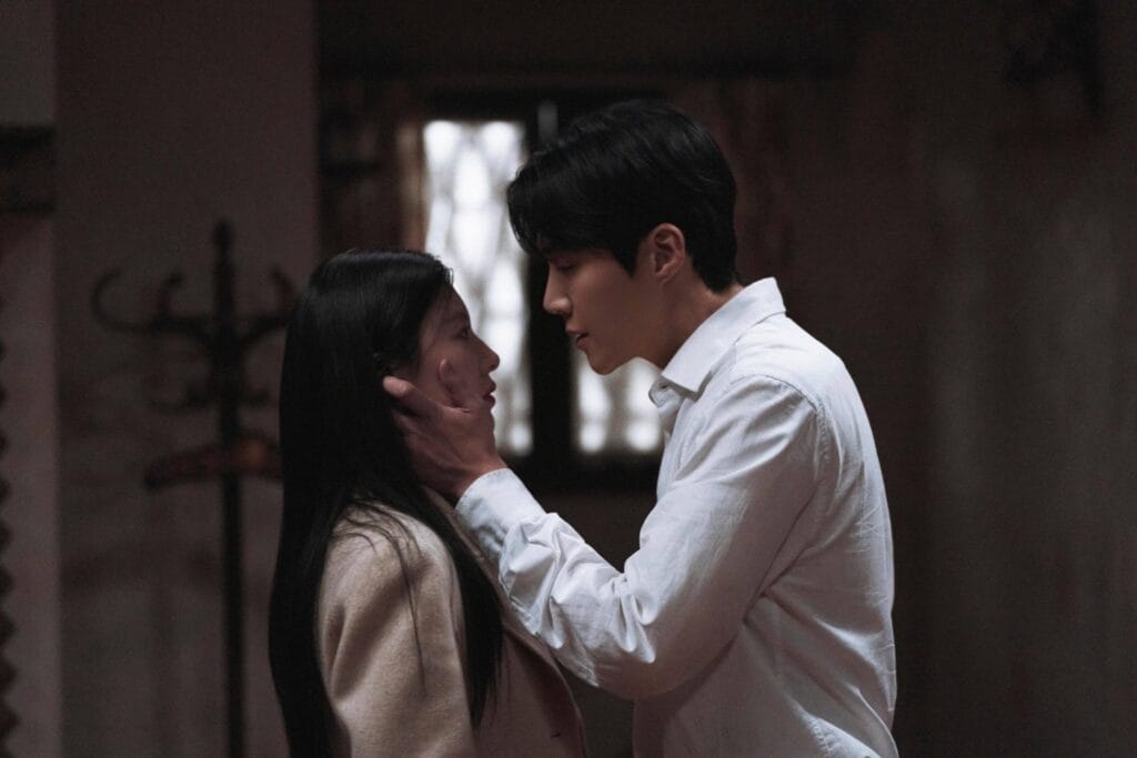 Kim Sun-Ho in a crisp white shirt gently cups a Go Youn-Jung ’s face with his hand as they stand inches apart in a dimly lit room. She gazes up at him, her expression soft and vulnerable, while he looks down at her with quiet intensity. Her long dark hair falls over her shoulder, and she wears a light beige coat. Behind them, a blurred window lets in faint natural light, casting gentle shadows across their faces.