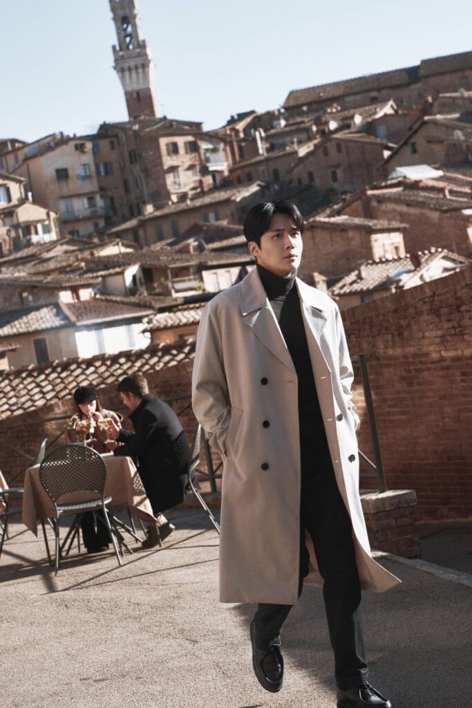 Kim Sun-Ho in a long beige trench coat over a black turtleneck and trousers walks with effortless grace along a sun-drenched rooftop terrace in a historic European city.
