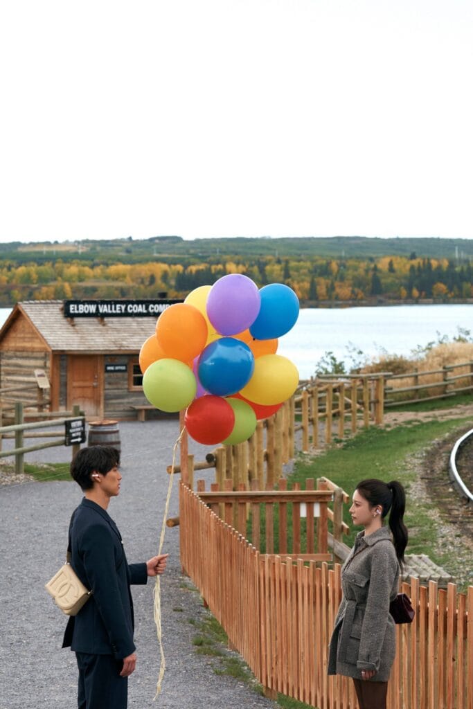 Go Youn-Jung and Sota Fukushi stand on opposite sides of a wooden fence at a rustic outdoor location, gazing at each other with quiet intensity. The man, dressed in a dark suit and carrying a beige crossbody bag, holds a cluster of colorful balloons — red, yellow, blue, green, and purple — as if offering them as a gesture or memory. The woman, wearing a textured gray coat with her hair in a ponytail, looks back at him with a thoughtful, slightly wistful expression. Behind them, a wooden cabin reads “ELBOW VALLEY COAL COMPANY,” and beyond it, autumn trees in golden hues line the shore of a calm lake under an overcast sky.