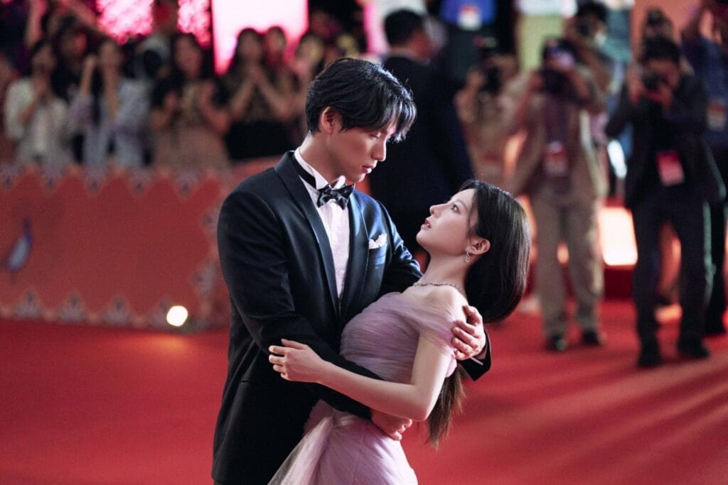 Sota Fukushi in a classic black tuxedo with bow tie gently embraces Go Youn-Jungin a soft lavender off-shoulder gown as they stand together on a red carpet. He holds her close, his gaze tenderly fixed on her, while she looks up at him with quiet affection. Behind them, photographers flash their cameras and fans applaud from behind barriers, capturing the magical moment under warm event lighting. Still image from the K-drama Can This Love Be Translated?