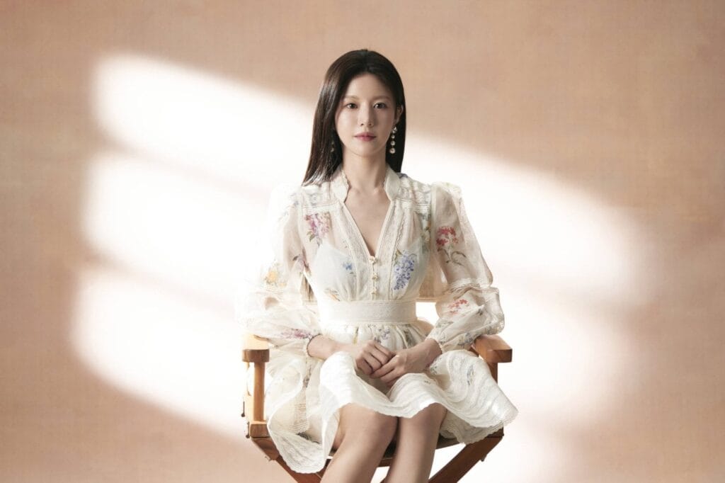 Go Youn-Jung with long dark hair sits gracefully on a wooden director’s chair, dressed in a delicate cream-colored dress adorned with floral embroidery and lace detailing. Her hands rest gently in her lap as she gazes calmly at the camera, exuding quiet poise and elegance.