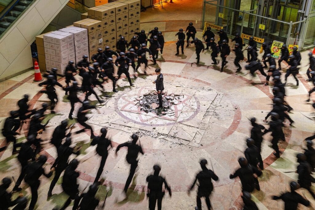 A lone man in a dark suit stands defiantly at the center of a shattered marble floor, surrounded by dozens of black-clad figures rushing toward him in a blur of motion. The scene is captured from above, emphasizing his isolation as he faces an overwhelming force. Behind him, stacks of cardboard boxes and an escalator suggest a modern indoor setting — perhaps a mall or corporate building. The floor beneath him is cracked open, revealing broken tiles and debris, with a faint circular symbol etched into the stone. The attackers, all wearing helmets and carrying batons, move with synchronized urgency, their blurred forms creating a sense of chaotic energy. Still image drom Cashero.