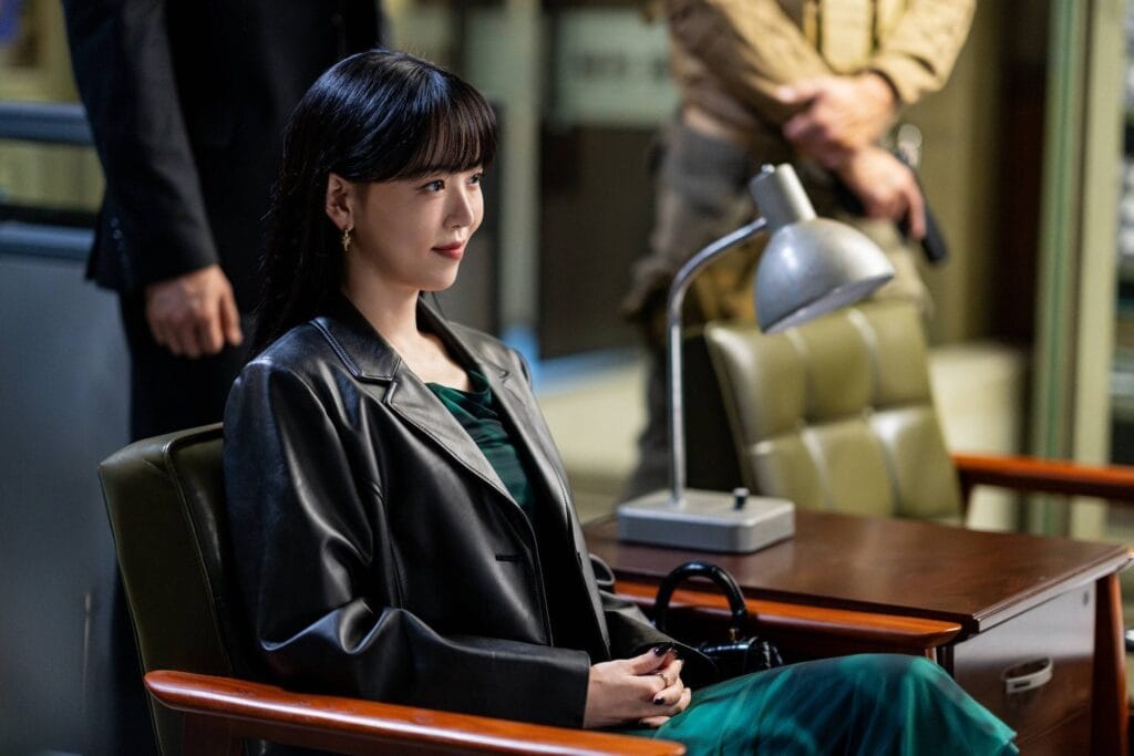 A woman with sleek dark hair and bangs sits poised in a leather chair, her expression calm yet subtly defiant as she gazes off-camera. She wears a tailored black leather blazer over a deep green dress, exuding quiet authority and style. Her hands are clasped gently in her lap, nails painted black, one finger adorned with a ring. Behind her, two men stand in the background — one in a dark suit, the other in tactical gear holding a firearm. Still image from Cashero.