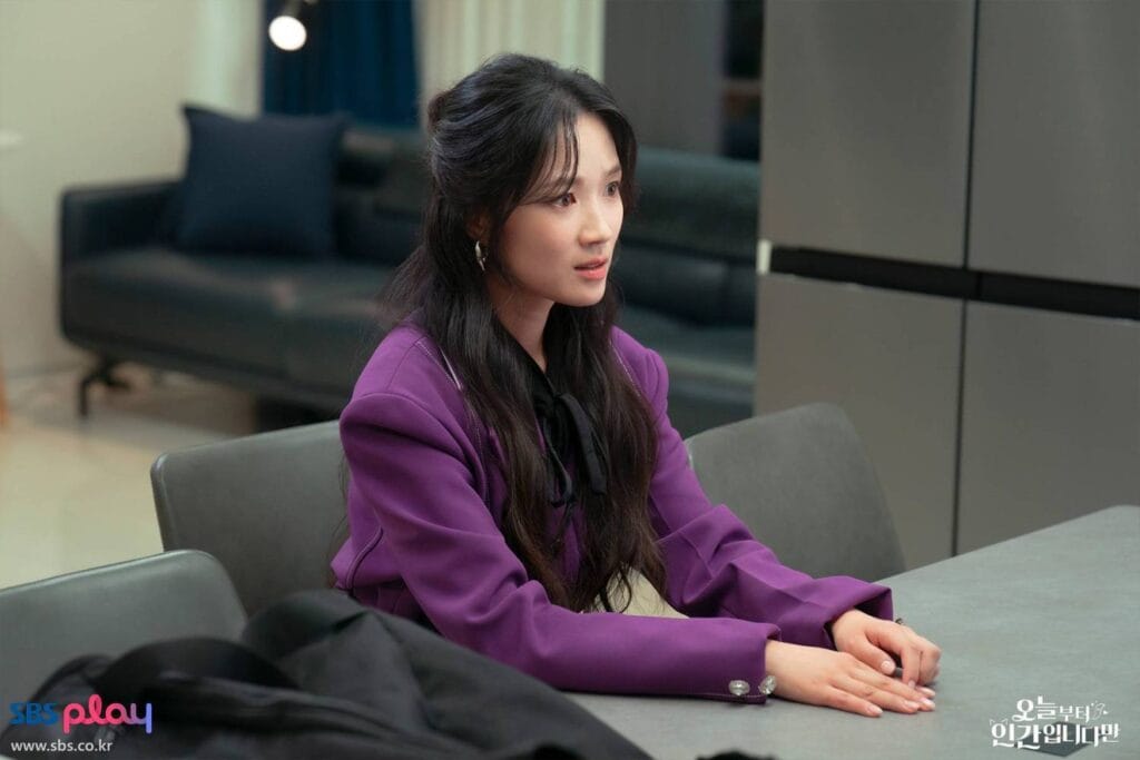 Kim Hye-yoon with long dark hair styled in a half-updo sits at a sleek gray conference table, her hands resting calmly on the surface. She wears a bold purple blazer over a black top with a bow detail at the neckline, exuding confidence and sophistication. Her gaze is directed off-camera with a thoughtful, slightly concerned expression — as if listening intently or contemplating a difficult decision. Behind her, a modern office or lounge space features a dark leather sofa, navy curtains, and minimalist decor. 