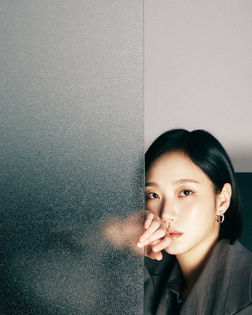 Kim Go Eun with  sleek dark hair and a contemplative gaze peers from behind a textured frosted glass panel, her fingers gently resting against it. She wears a tailored gray blazer over a dark top, exuding quiet confidence and understated sophistication. A delicate hoop earring catches the light near her ear. The background is split — one side features the cool, grainy texture of the translucent glass; the other, a smooth matte gray wall — creating a visual contrast between concealment and revelation. Soft, directional lighting highlights her features, emphasizing her calm yet intense expression.