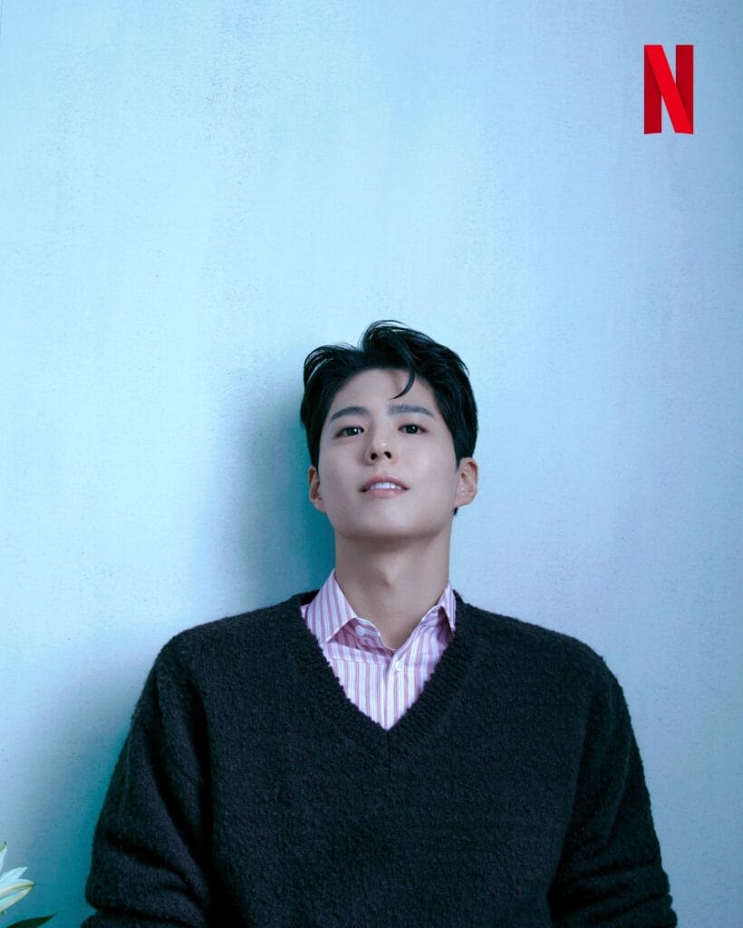 Park Bo-gum with neatly styled dark hair leans casually against a soft light-blue textured wall, gazing thoughtfully upward. He wears a cozy black V-neck sweater over a pink-and-white striped collared shirt, creating a relaxed yet polished look.