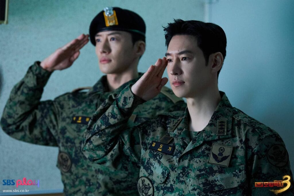 Two men in South Korean military uniforms stand side-by-side, saluting with serious, focused expressions. The man in the foreground wears a digital camouflage uniform with name tag “김도기 / KIM” and airborne insignia on his chest; his gaze is steady and resolute. Behind him, another soldier in a similar uniform and black beret mirrors the salute, his face calm but intense. 