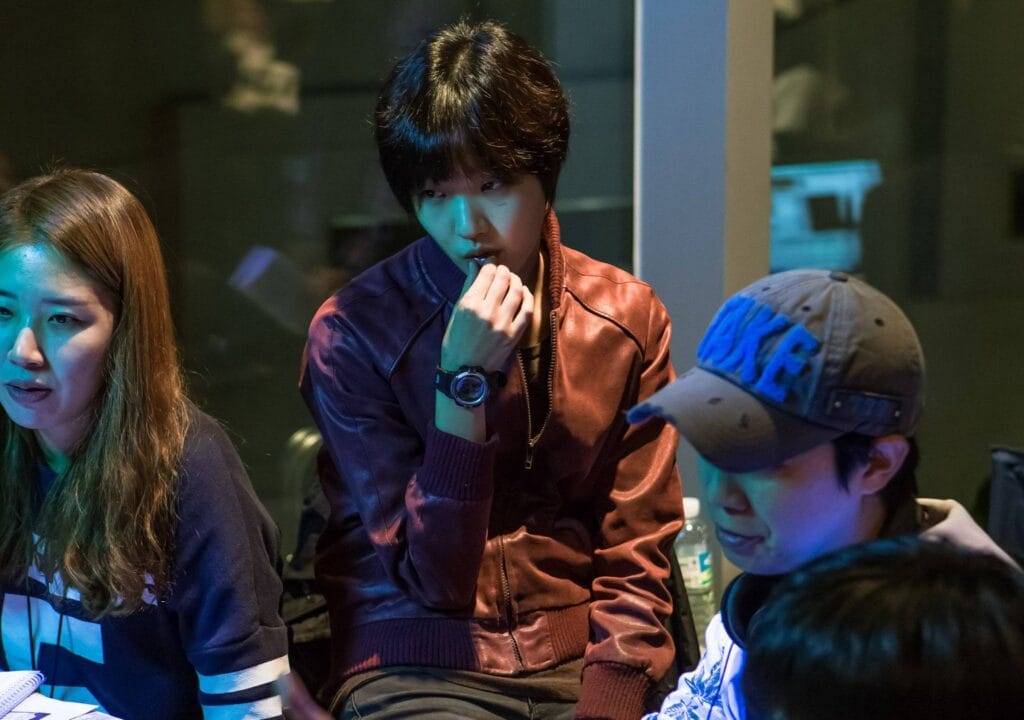 Kim Go Eun with short dark hair wears a two-tone brown-and-purple leather bomber jacket and rests their chin on their fist, eyes narrowed in concentration—wearing a prominent black watch attends  a conversation.