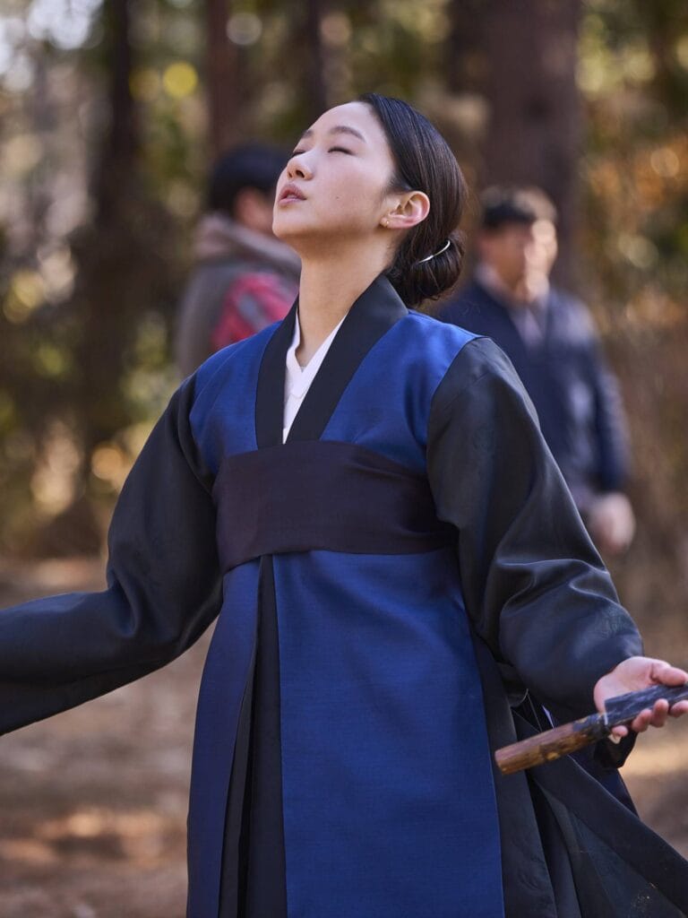 Kim Go-eun  in traditional Korean hanbok stands in a sun-dappled forest, her eyes closed and face lifted toward the sky in a moment of quiet contemplation or resolve. She wears a deep blue jeogori with black sleeves and a wide black sash, her dark hair neatly pinned back with a simple metal hairpin. In her right hand, she holds the wooden hilt of a sword.