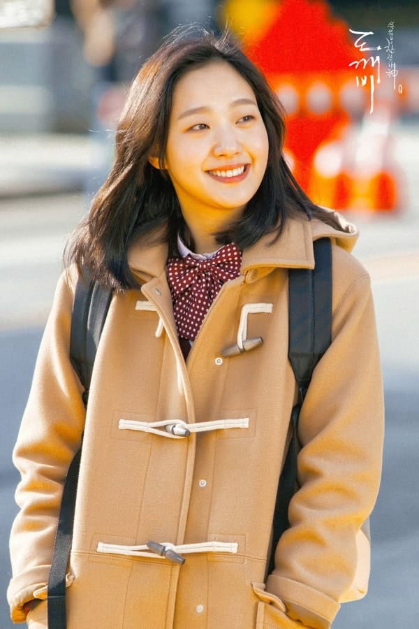 Kim Go   Eun with shoulder-length dark hair smiles brightly as she walks outdoors, her face lit by soft golden sunlight. She wears a cozy beige duffle coat with white toggle closures, a red-and-white polka-dot bow tie, and carries a black backpack over her shoulders. Her expression is cheerful and carefree — eyes crinkled in genuine delight.