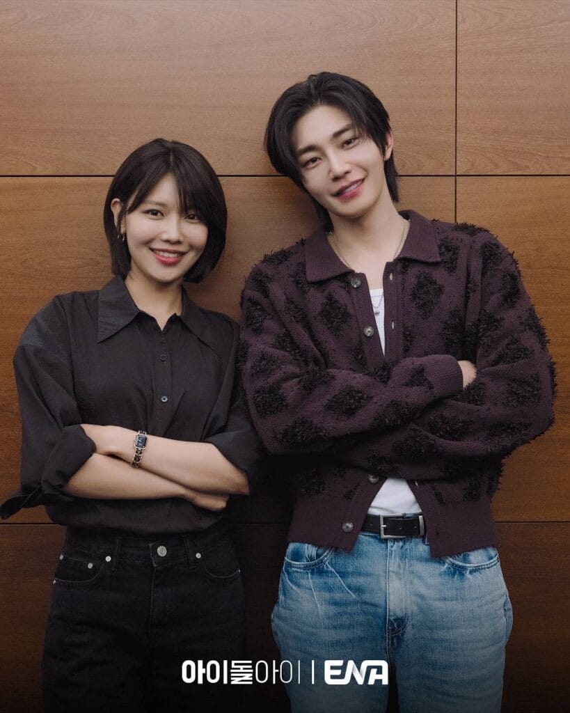 Actors Choi Soo-young and Kim Jae-young pose side-by-side against a warm wooden-paneled wall, smiling confidently at the camera for a promotional photo shoot  of Idol I.