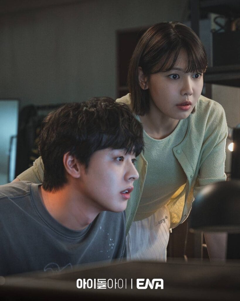 A young man and woman lean closely over a computer screen in a dimly lit room, their expressions locked in shared concentration. The man, in a gray T-shirt, sits slightly in front, mouth slightly open as if reacting in real time. Behind him, the woman—wearing a soft mint-green cardigan over a striped top—leans forward, eyes wide and alert, her hand resting lightly on his shoulder. Warm light from the screen illuminates their faces, casting subtle shadows that heighten the sense of suspense. 