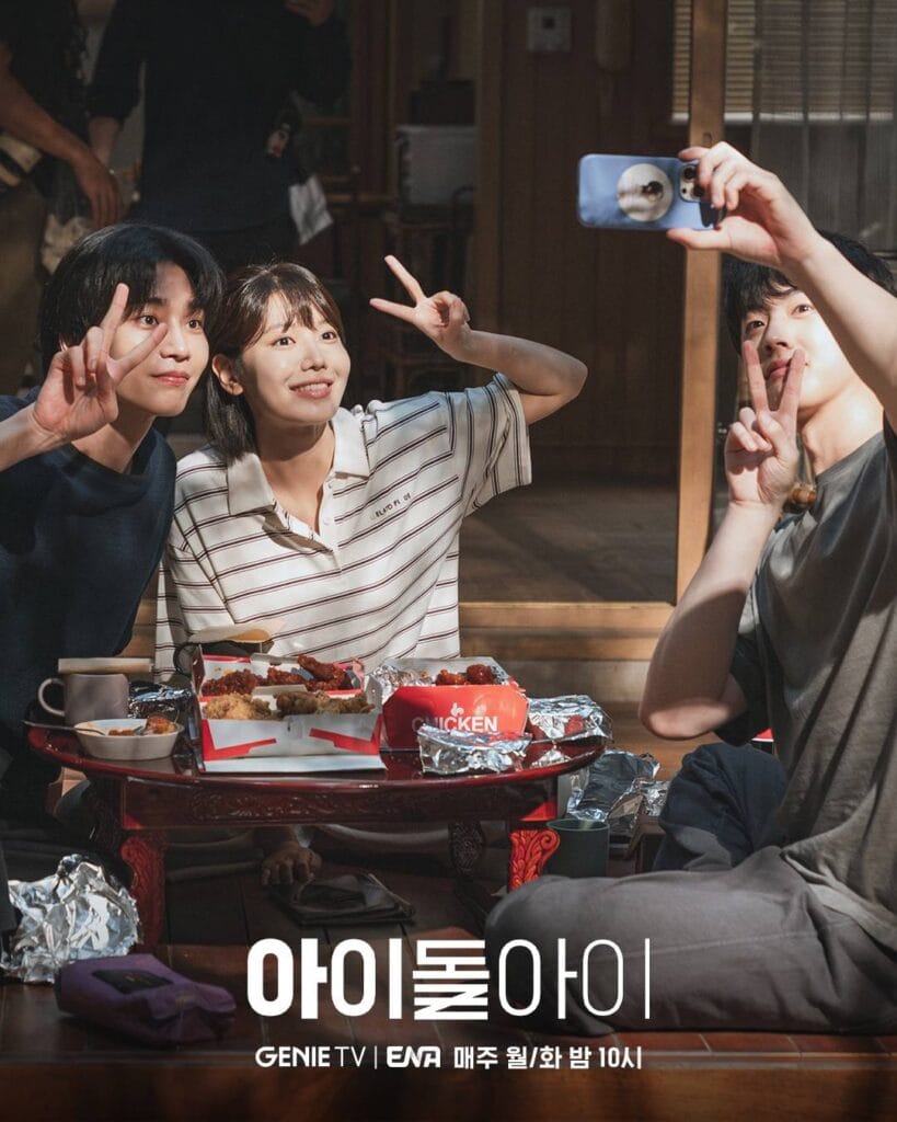 Three persons pose joyfully for a selfie during a casual meal break on set. The woman in the center—wearing a striped polo shirt—smiles brightly while making a peace sign with both hands. Flanking her, two men also flash peace signs: one on the left in a navy sweater, the other on the right holding up a blue smartphone to take the photo. 