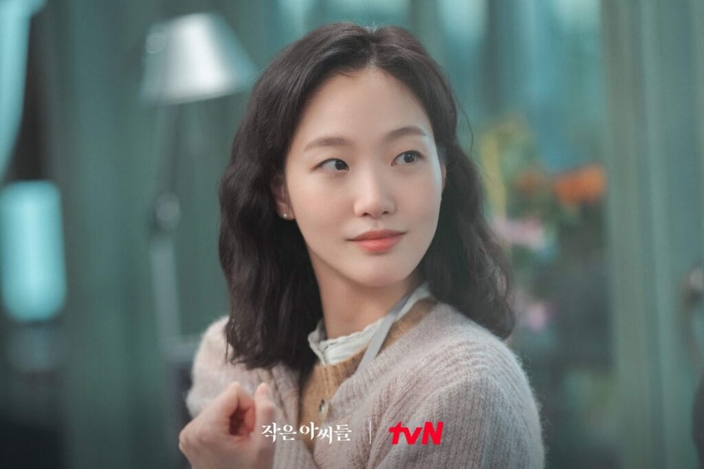 Kim  Go  Eun with shoulder-length wavy dark hair gazes thoughtfully to her right, a subtle, warm smile playing on her lips. She wears a cozy light-pink knit cardigan over a cream blouse with delicate lace trim, and small pearl earrings glint softly in the ambient light. Her hands are gently clasped near her chest.