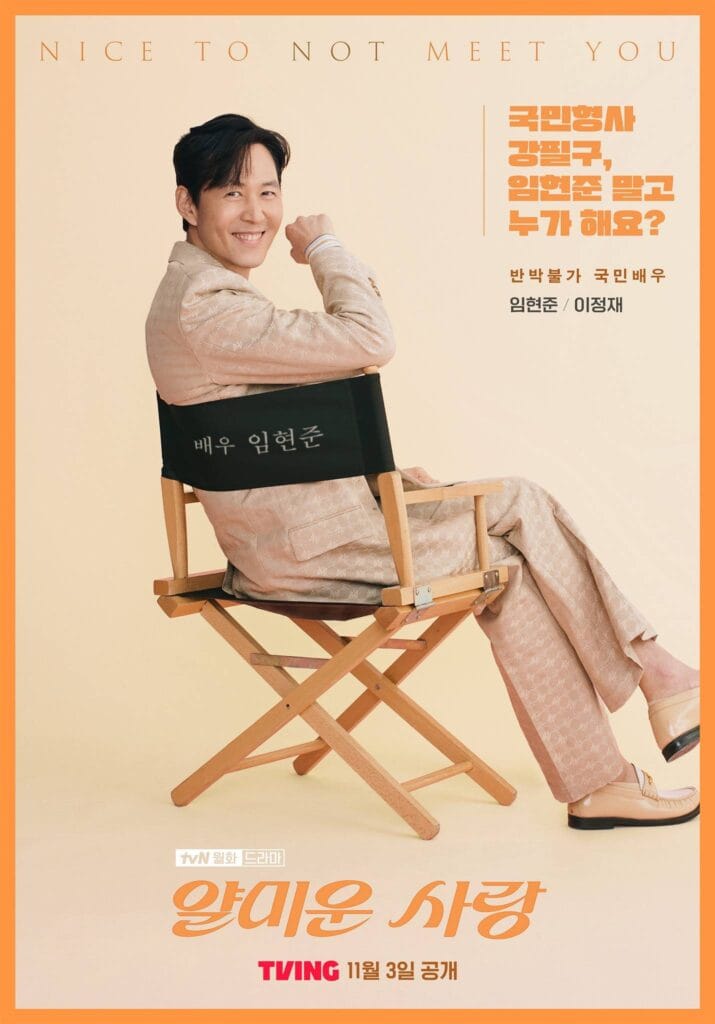 A smiling man sits confidently in a wooden director’s chair against a warm cream background, wearing a coordinated beige patterned suit with matching loafers. He leans back with one arm resting on the chair’s back, his expression playful and charismatic. 