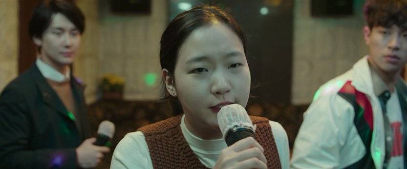 Kim  Go  Eun with short dark hair sings into a handheld microphone, her expression deeply focused and tender—lips parted mid-phrase, eyes slightly downcast. She wears a white turtleneck under a textured brown knit vest, and soft stage lighting casts a gentle glow on her face. Behind her, two men stand slightly out of focus: one in a dark suit holding a mic, the other in a white jacket with red-and-green trim, both watching her with quiet attention. 