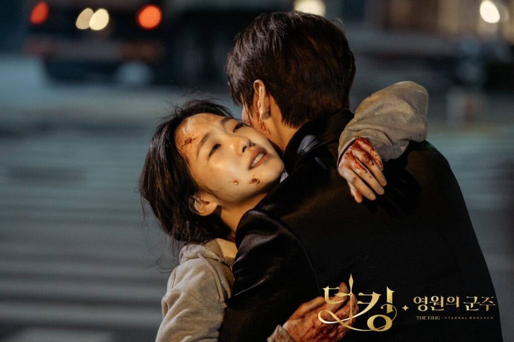 Kim  Go Eun with blood smudged on her forehead and cheek clings desperately to a man in a dark coat, her arms wrapped tightly around his neck. Her face is tilted upward, eyes glistening with tears — part relief, part sorrow — as she presses her cheek against his shoulder.