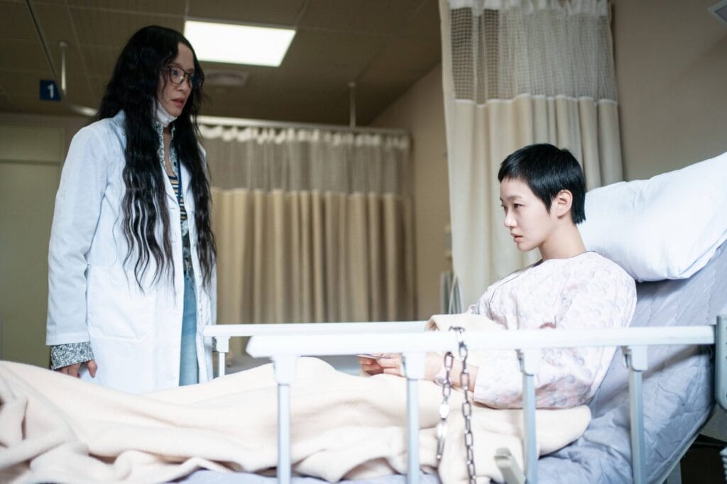 Kim Go Eun  with a short pixie haircut, wears a pale floral hospital gown and is restrained by a metal chain attached to the bed rail. while a woman with long dark hair and glasses—wearing a white lab coat stands beside the bed.