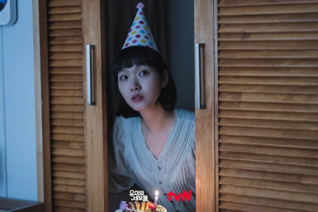Kim  Go Eun with short dark hair and bangs peeks out from between two wooden closet doors, wearing a colorful polka-dotted party hat. She holds a small birthday cake with one lit candle, her expression a mix of hope, vulnerability, and quiet loneliness. She wears a soft cream-colored lace-trimmed blouse, and the dim lighting casts gentle shadows across her face. 