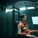 Kim Go-eun with short dark hair sits in a high-tech interrogation chair inside a sterile, dimly lit room with tiled walls and overhead fluorescent lighting. She wears an orange jumpsuit with a yellow identification tag on her chest and has a blood pressure cuff on her left arm. Her right hand holds a response device, and she stares intently at a monitor displaying six Korean questions—such as “Is your name [redacted]?” and “Have you ever met An Won-su at the detention center?”—suggesting a polygraph or cognitive test. Behind her looms a large metal archway, reinforcing the clinical, oppressive atmosphere.